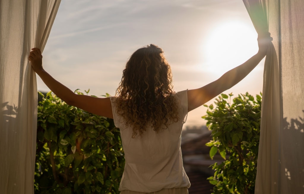 Woman waking up in the morning and looking at the sunrise through the window